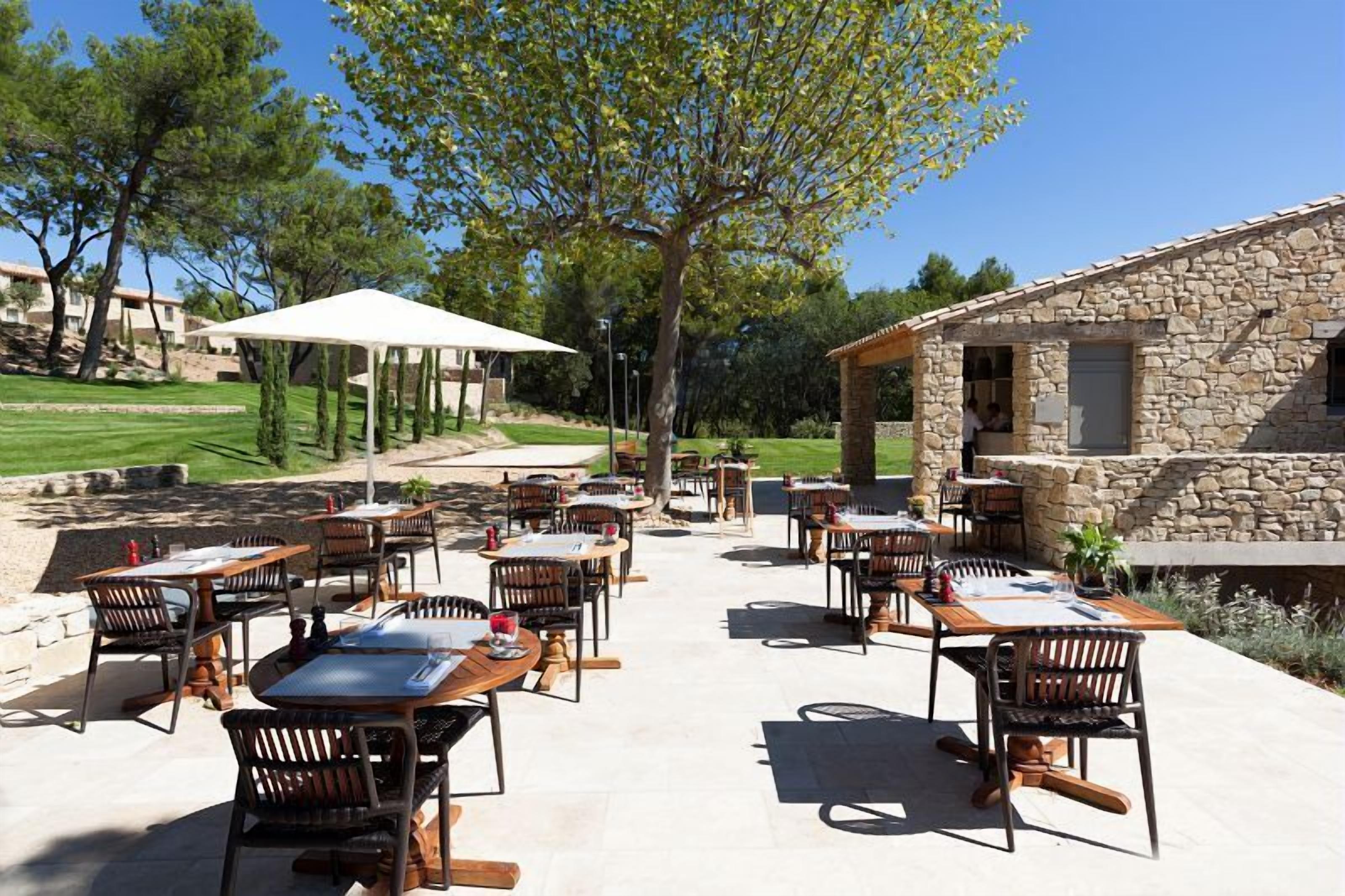 Une terrasse extérieure avec plusieurs tables et chaises, un bâtiment en pierre et un grand arbre qui offre de l’ombre.