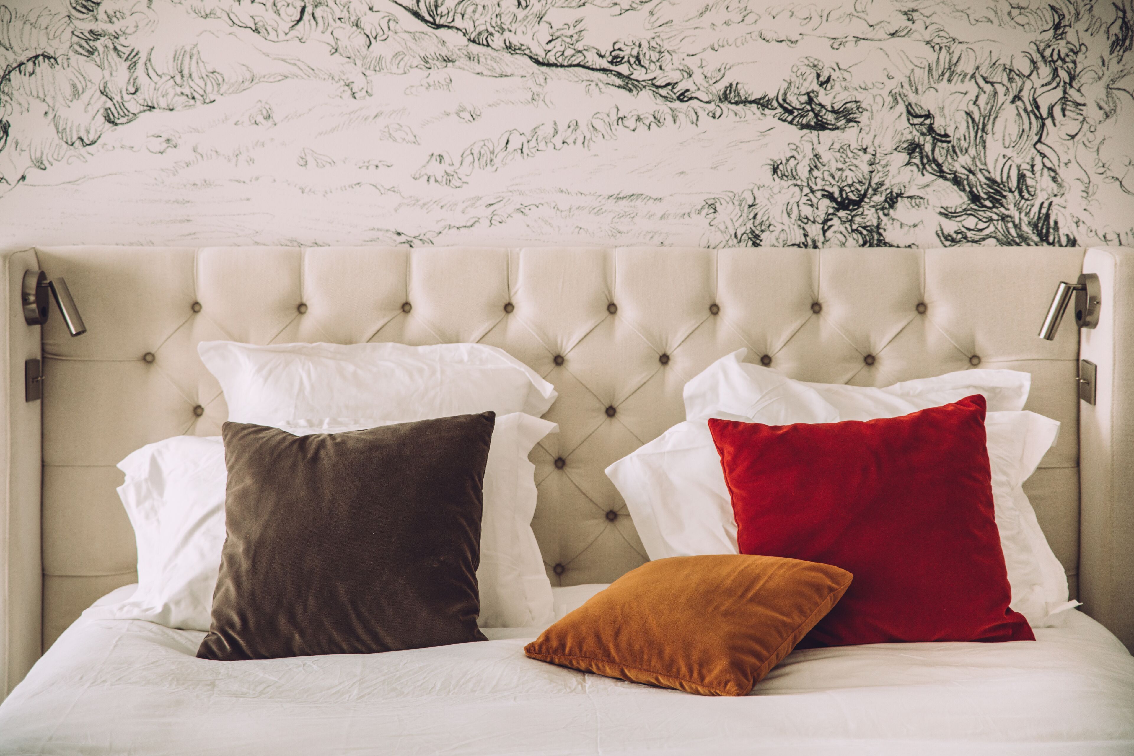 A bed with a tufted headboard, white bedding, and three pillows in brown, red, and orange.