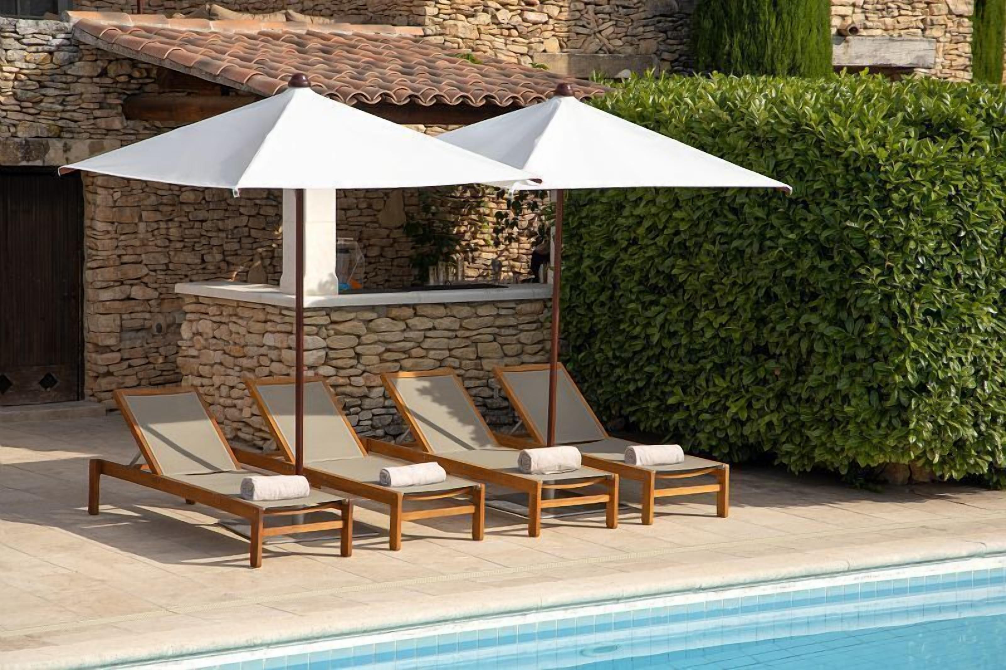 Ein Poolbereich mit Liegestühlen und Sonnenschirmen, einer Steinmauer und einem ordentlich geschnittenen Heckenstreifen.