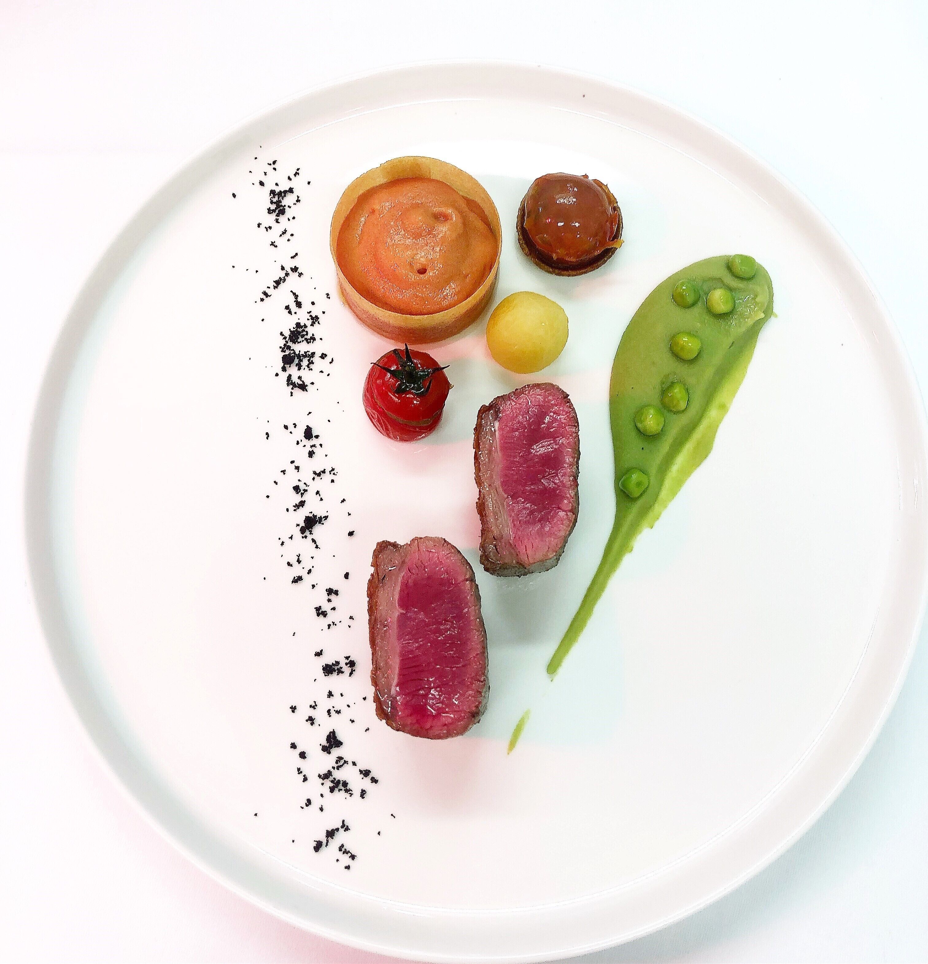 A plate with various food items including a round pastry, a slice of meat, a pea pod, a cherry tomato, and a small yellow item, all arranged on a white plate.