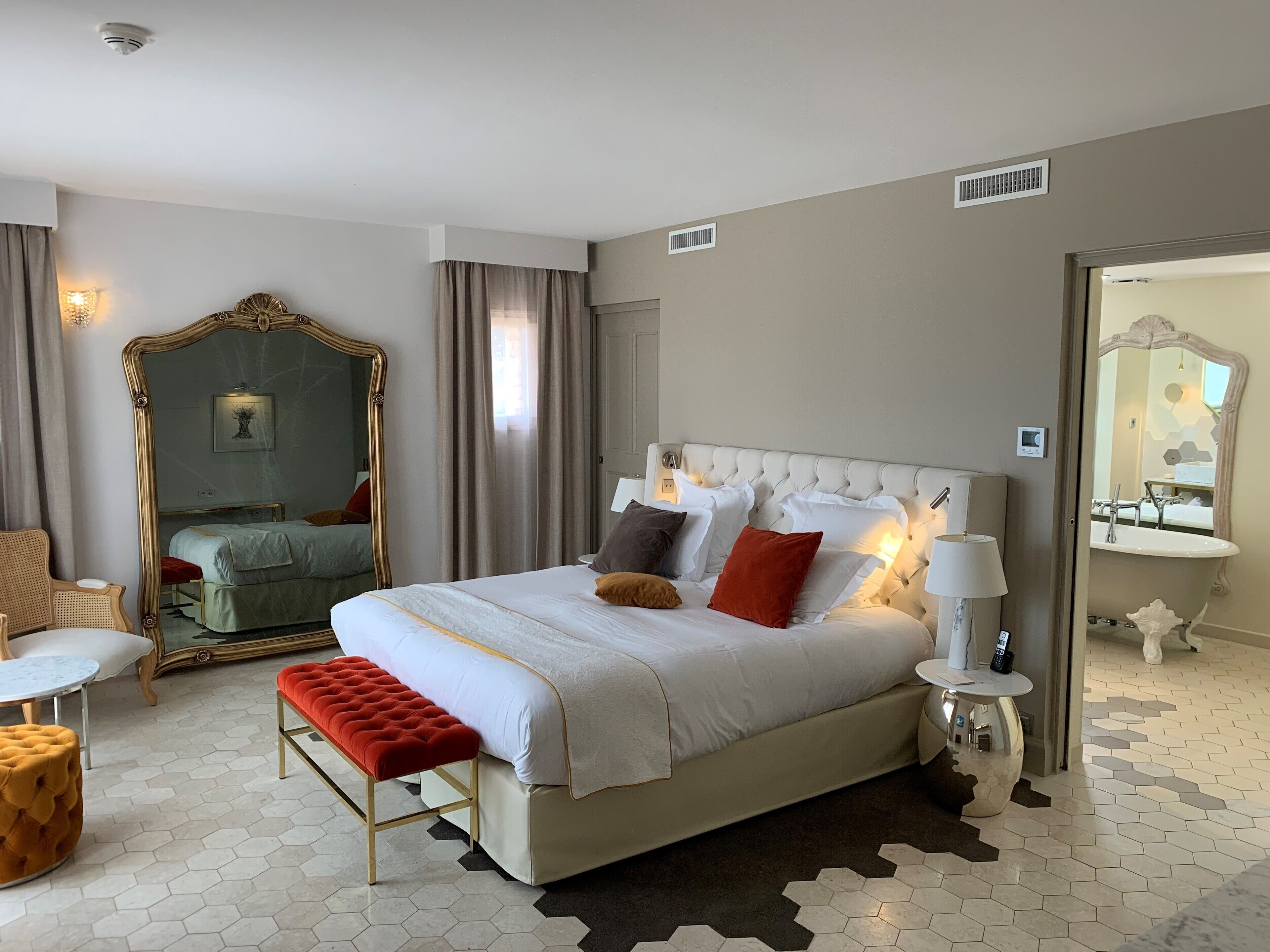A hotel room with a large bed, a round table, a red bench, a mirror, and a bathroom visible through an open door.