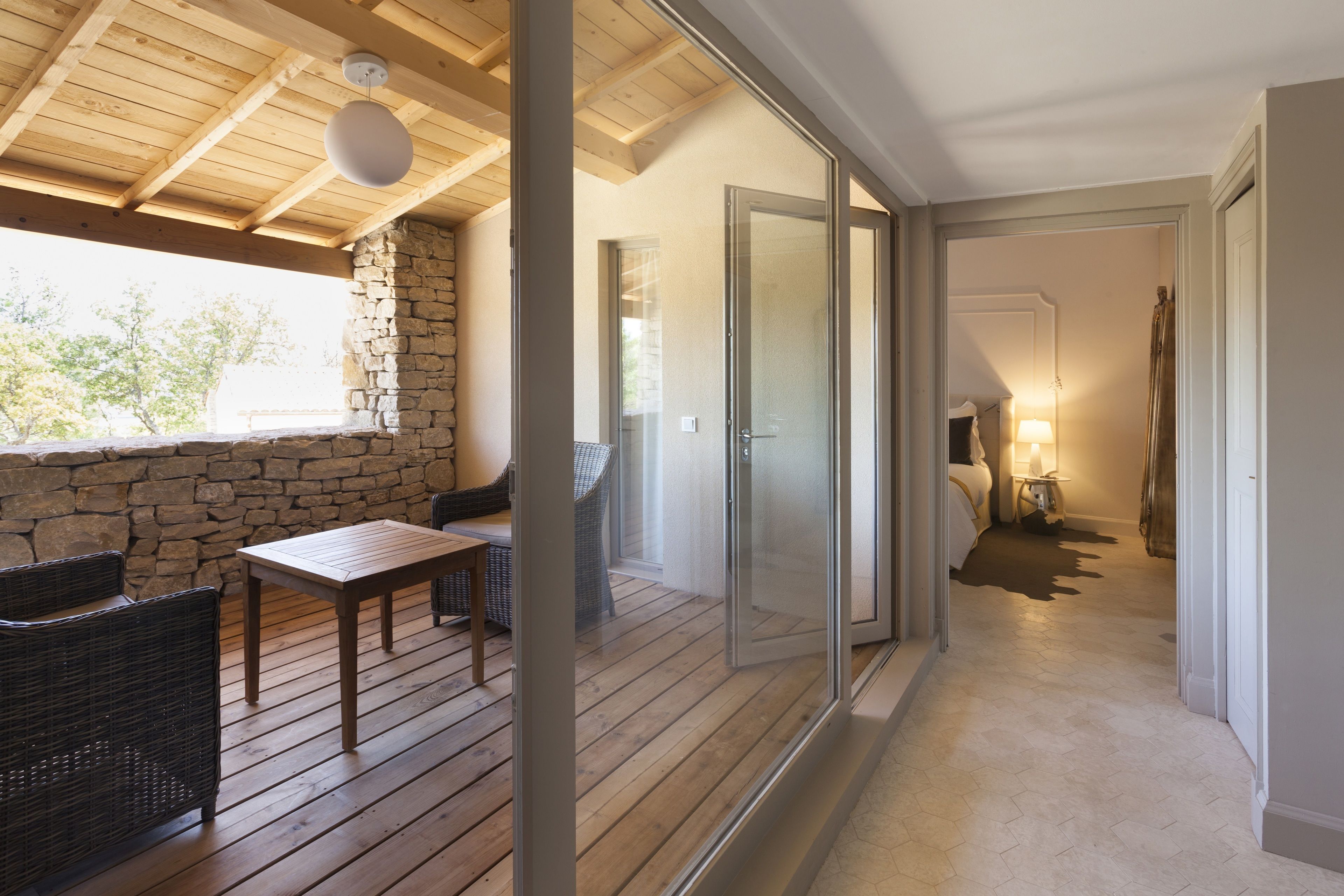 A wooden deck with wicker furniture, a stone wall, and a glass door leading to a bedroom with a bed, nightstand, and lamp.