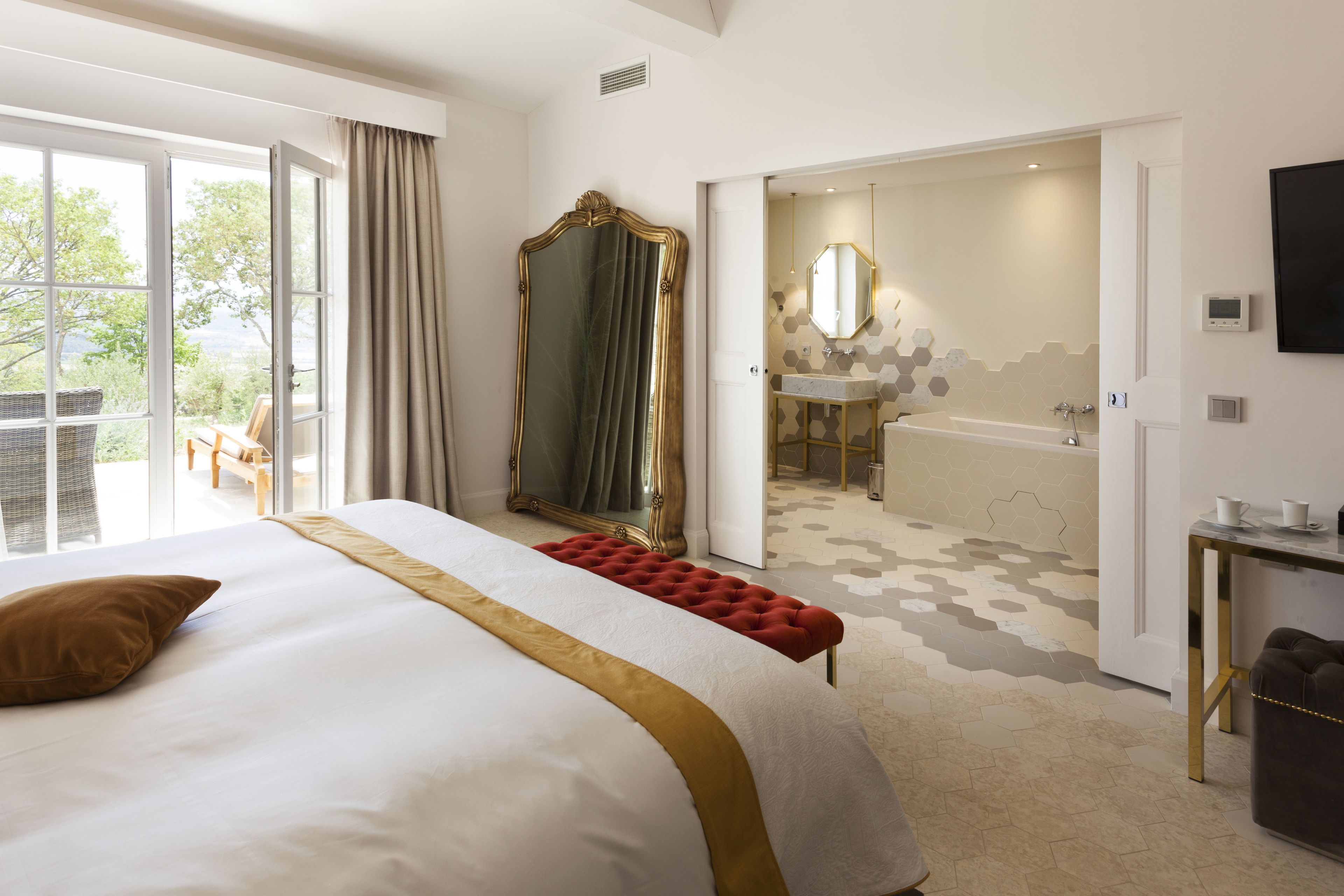 A hotel room with a large bed, a red ottoman, a large mirror, a bathroom with a bathtub, and a view of the outdoors.