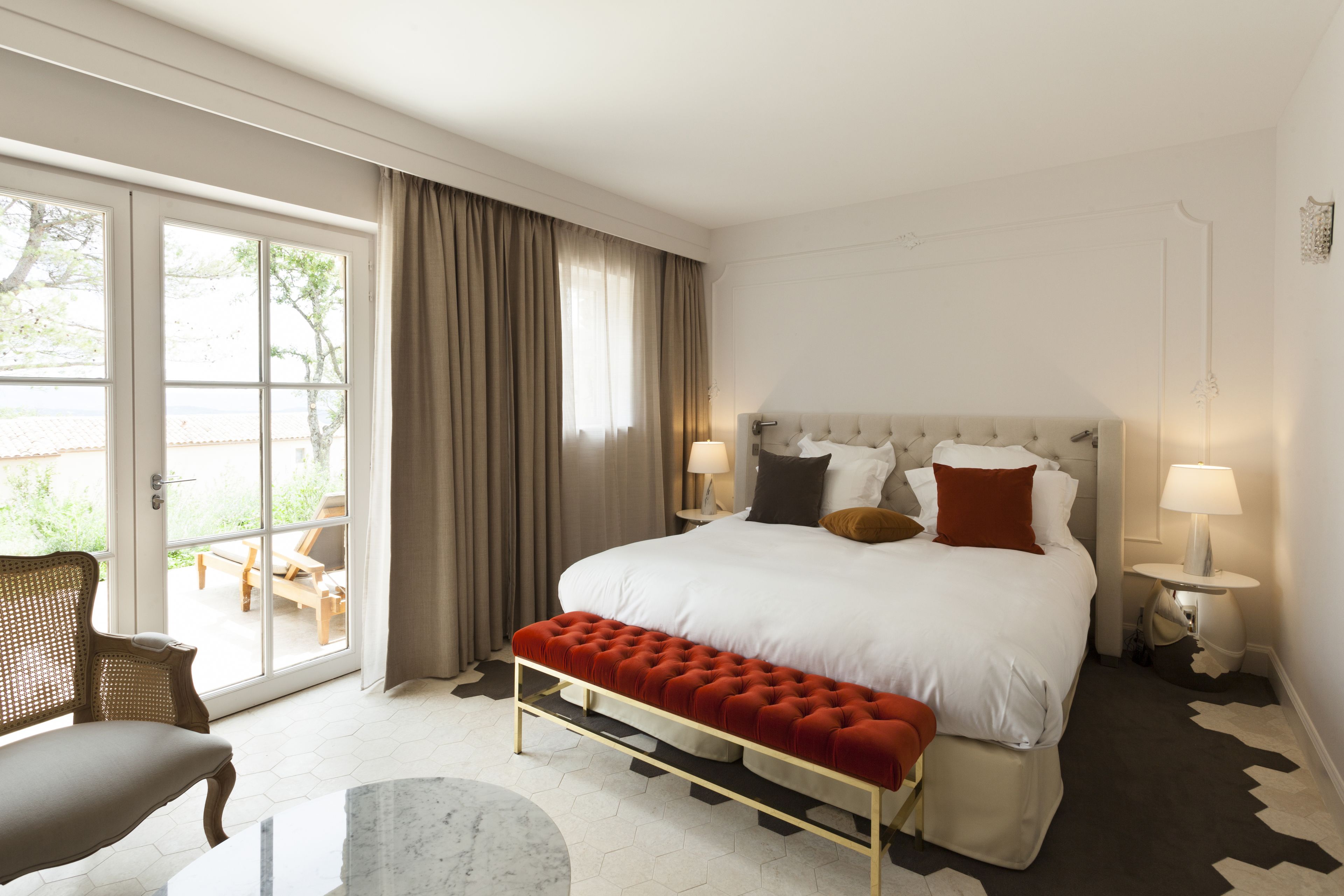 A hotel room with a large bed, a red bench, a chair, a round table, and a view through a glass door.