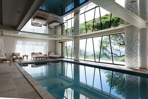 Une piscine intérieure offrant une vue dégagée sur la verdure environnante, de grandes fenêtres et un espace détente avec des chaises et une table.