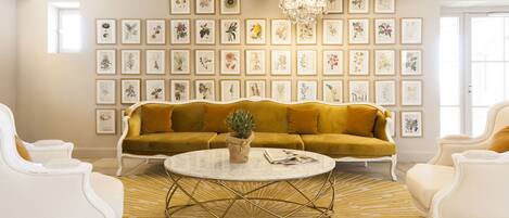 A living room with a mustard yellow sofa, white armchairs, a round coffee table, and a wall of framed botanical illustrations.
