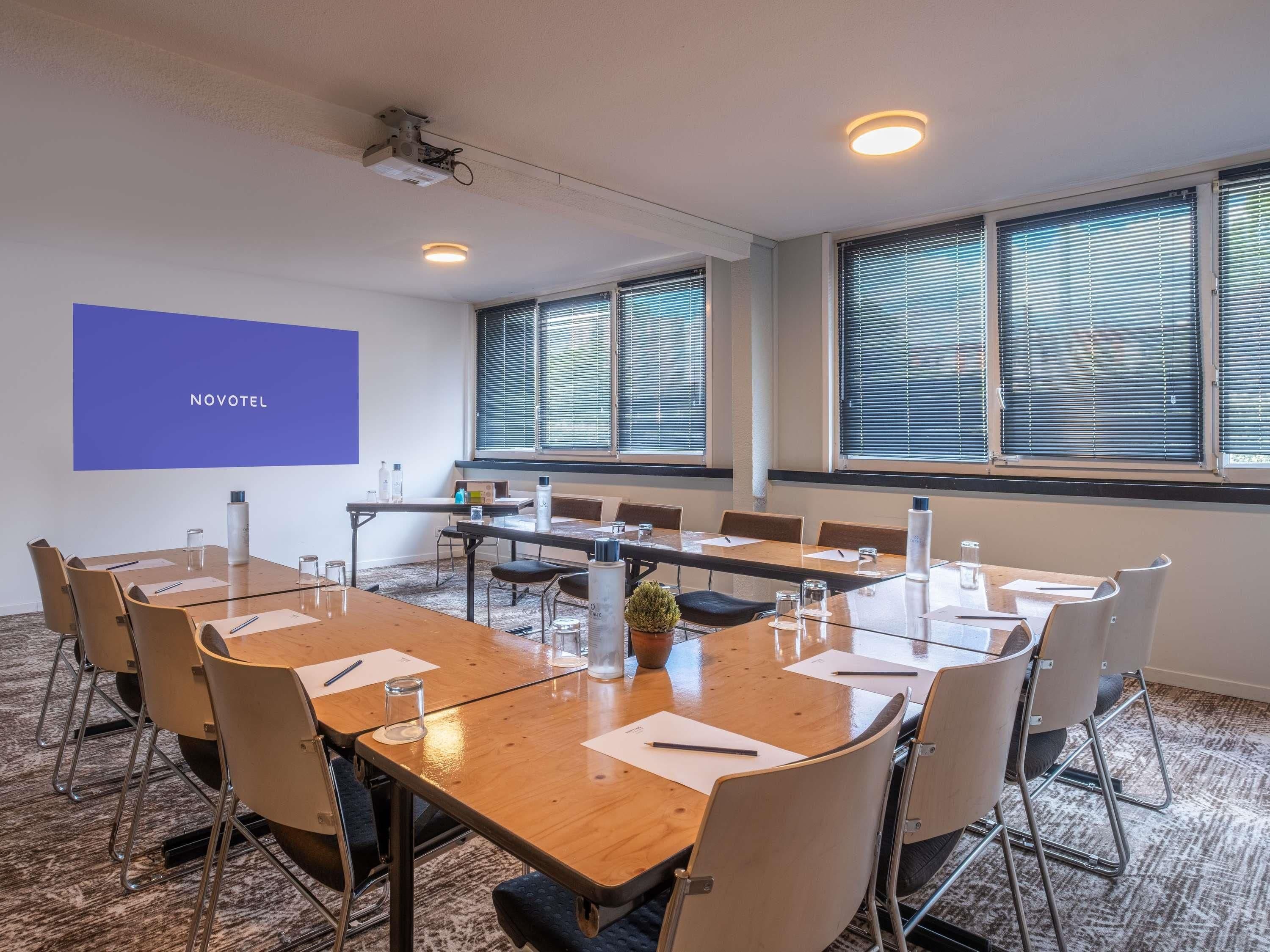 Een vergaderzaal met een lange tafel, stoelen, een projectorscherm en grote ramen met zonwering.