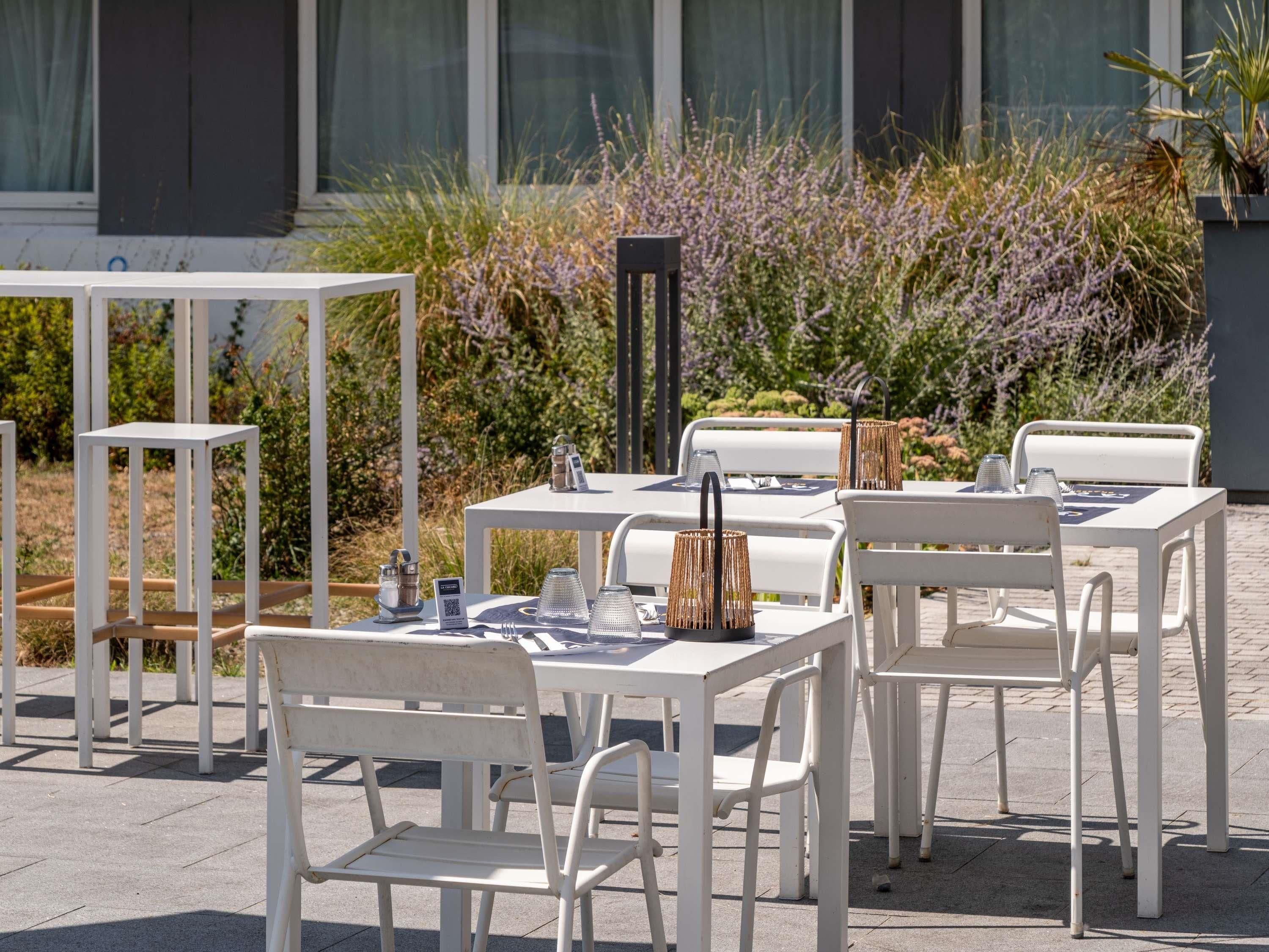 An outdoor seating area with white tables and chairs, surrounded by plants and a building in the background.