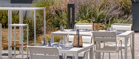An outdoor seating area with white tables and chairs, surrounded by plants and a building in the background.