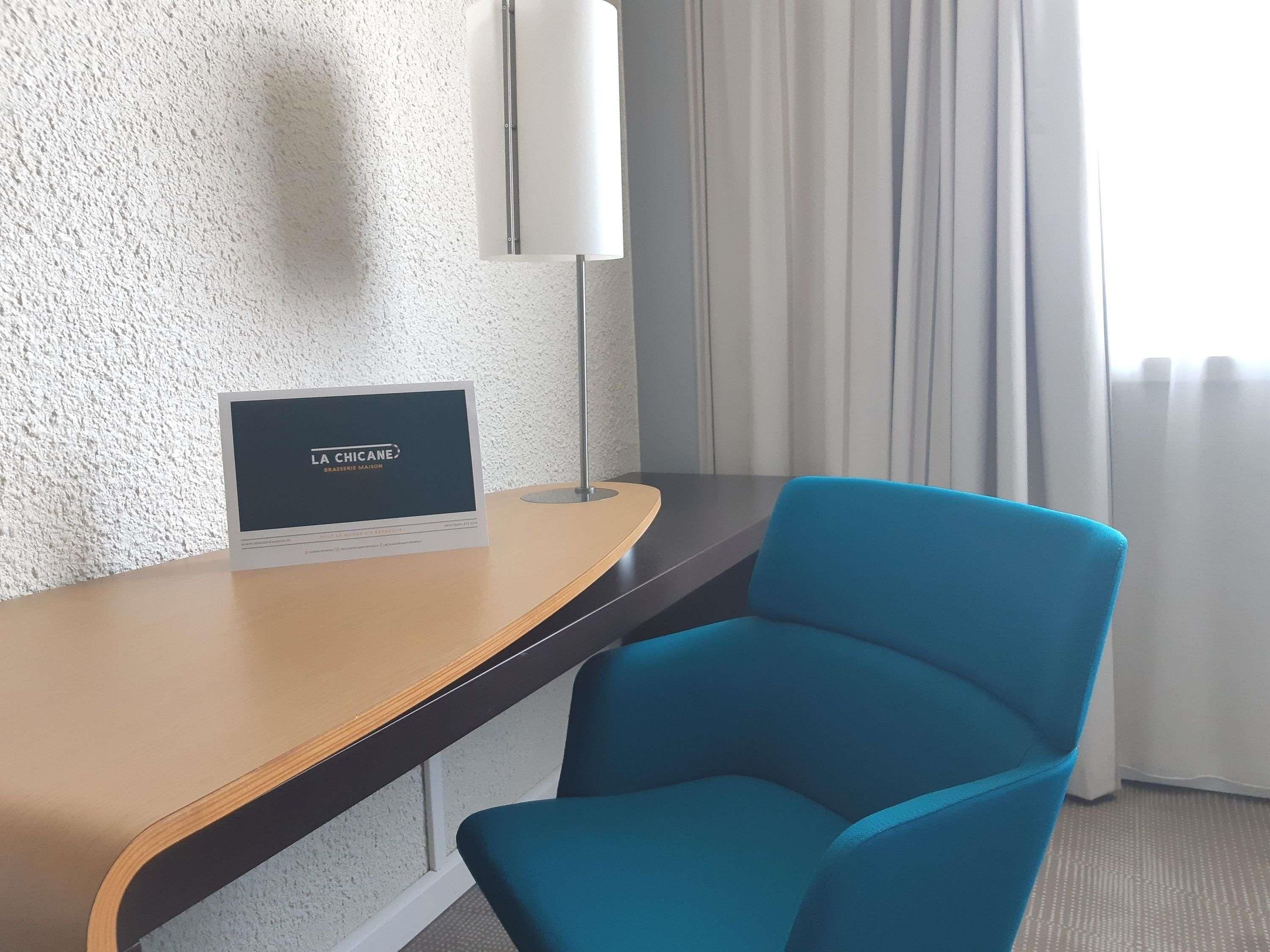 Una habitación de hotel con un escritorio de madera, una silla azul, una lámpara de pared y un letrero que dice 'LA CHICANE BRASSERIE MAISON'.