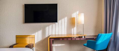 Habitación de hotel con cama, escritorio, silla, televisor de pantalla plana, lámpara y cortinas.