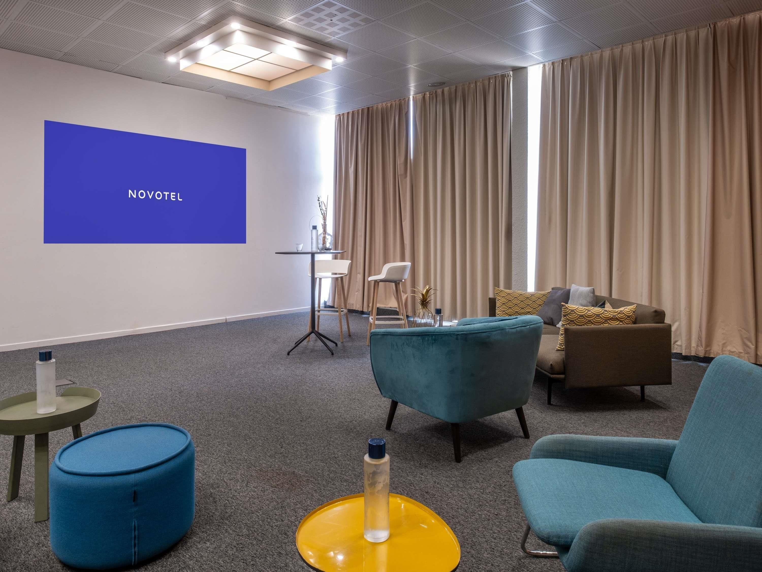 A modern hotel room with a large screen displaying 'NOVOTEL,' a cozy seating area with a sofa and chairs, a small round table, and a blue ottoman.