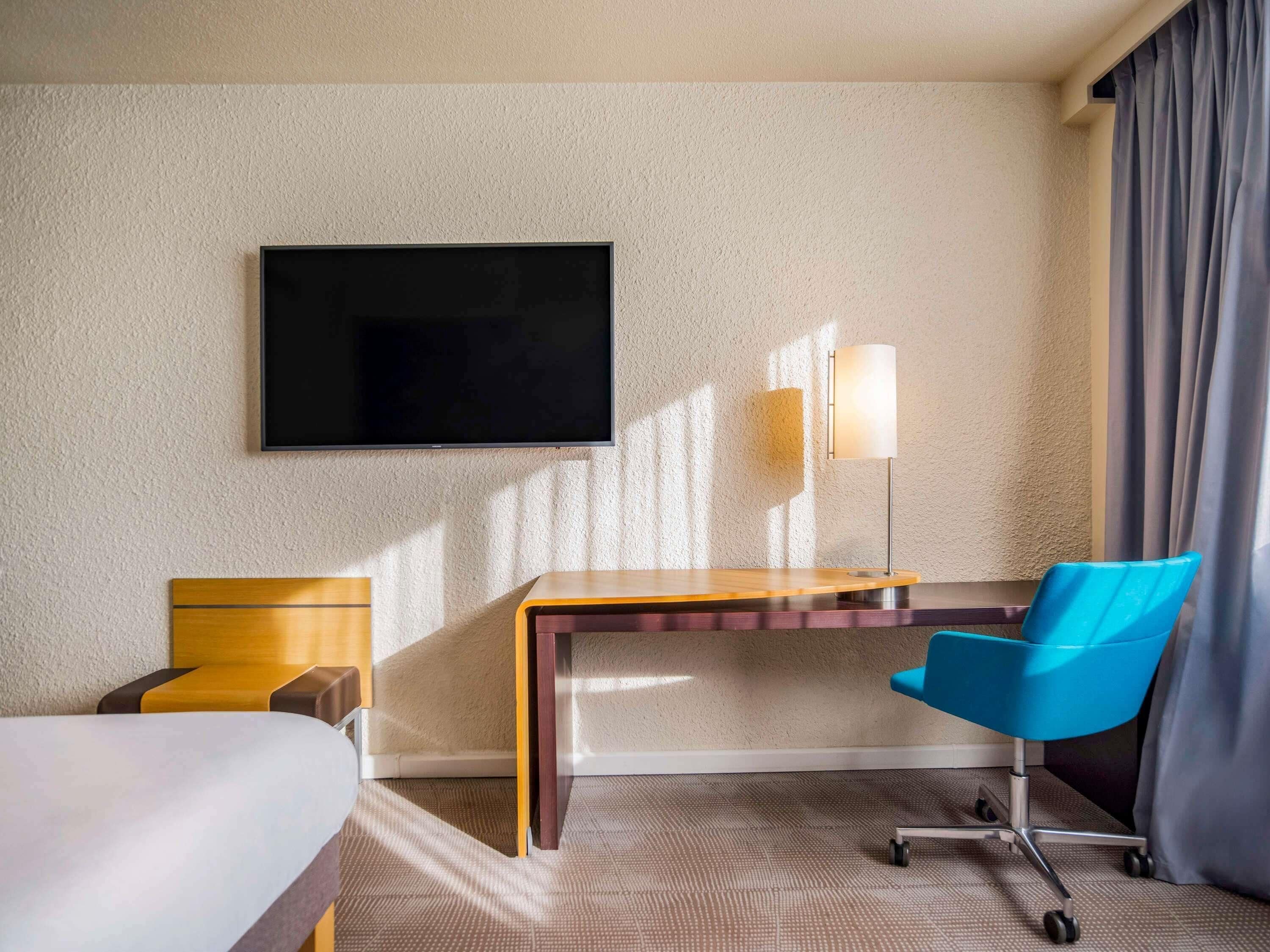 A hotel room with a bed, desk, chair, flat-screen TV, lamp, and curtains.
