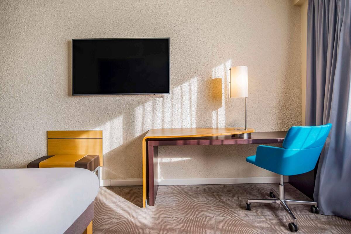 Une chambre d’hôtel équipée d’un lit, d’un bureau, d’une chaise, d’un téléviseur à écran plat, d’une lampe et de rideaux.