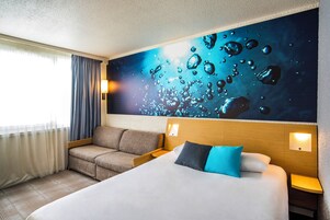 A hotel room with a large bed, a sofa, a wall mural, and a window with curtains.
