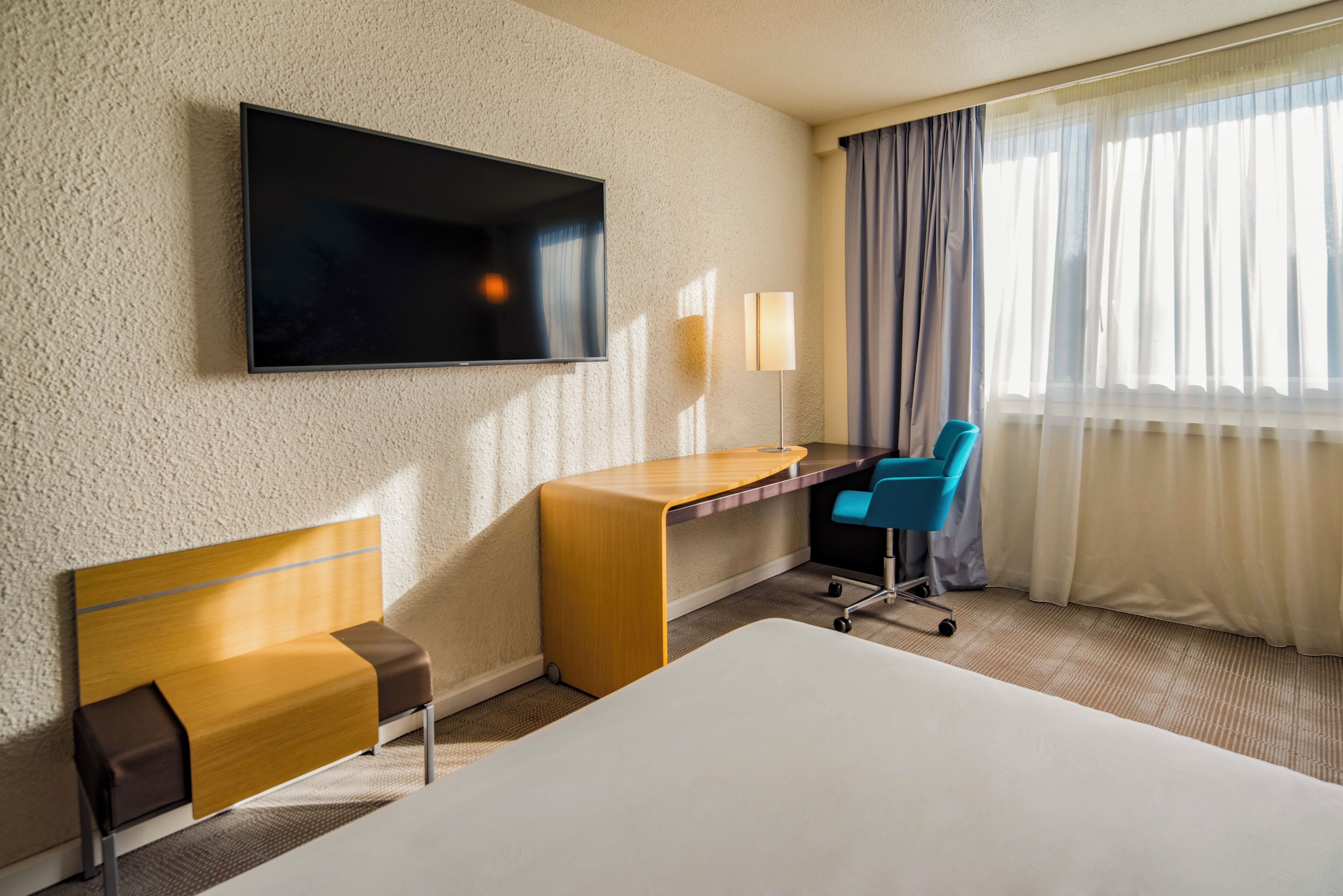 A hotel room with a bed, a desk, a chair, a TV, and a window with curtains.