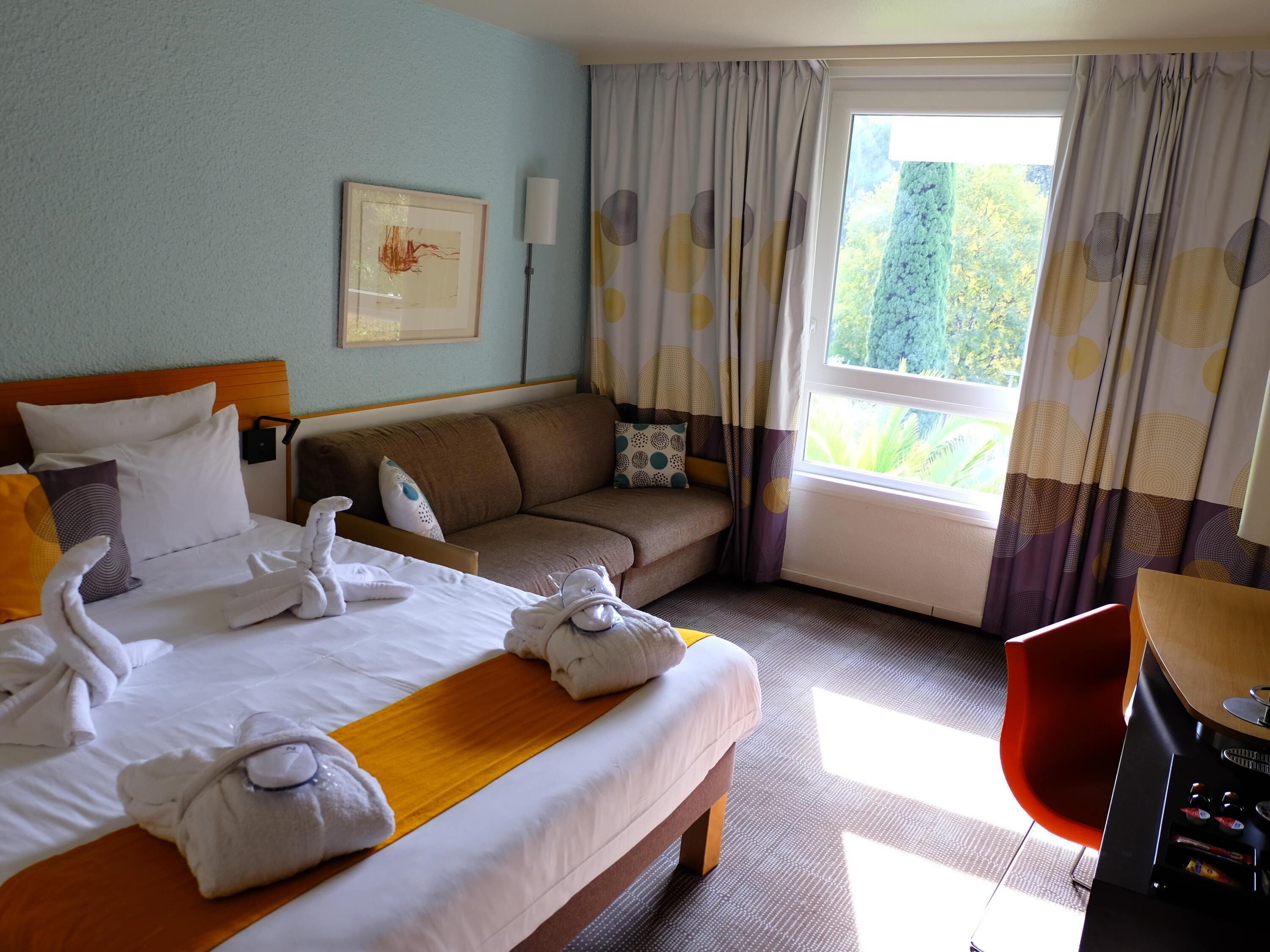 Una habitación de hotel con cama, sofá, mesa de comedor y ventana con cortinas.