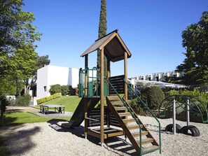 A playground with a wooden slide and stairs, surrounded by gravel and greenery.