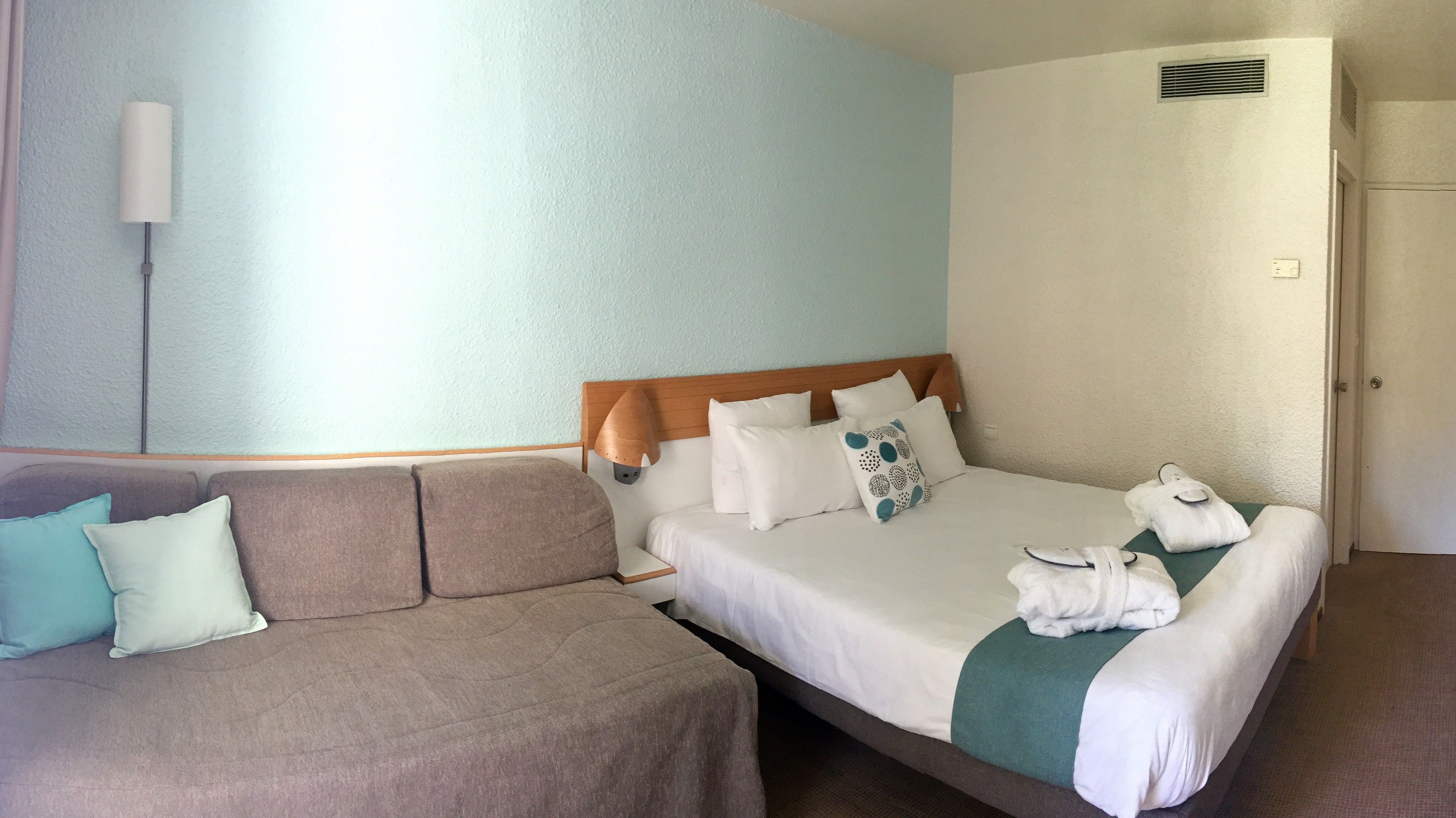 A hotel room with a bed, a sofa, and a wall-mounted lamp.