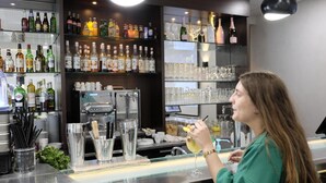 Uma área de bar com uma máquina de café, prateleiras de garrafas de bebidas e diversos copos.