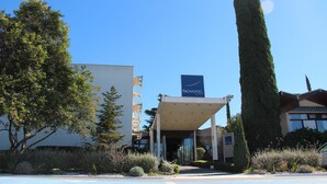Entrada de hotel com céu claro, placa da Novotel Hotels e área de estacionamento.