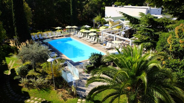 Área de piscina com espreguiçadeiras e guarda-sóis, rodeada por vegetação e um edifício ao fundo.