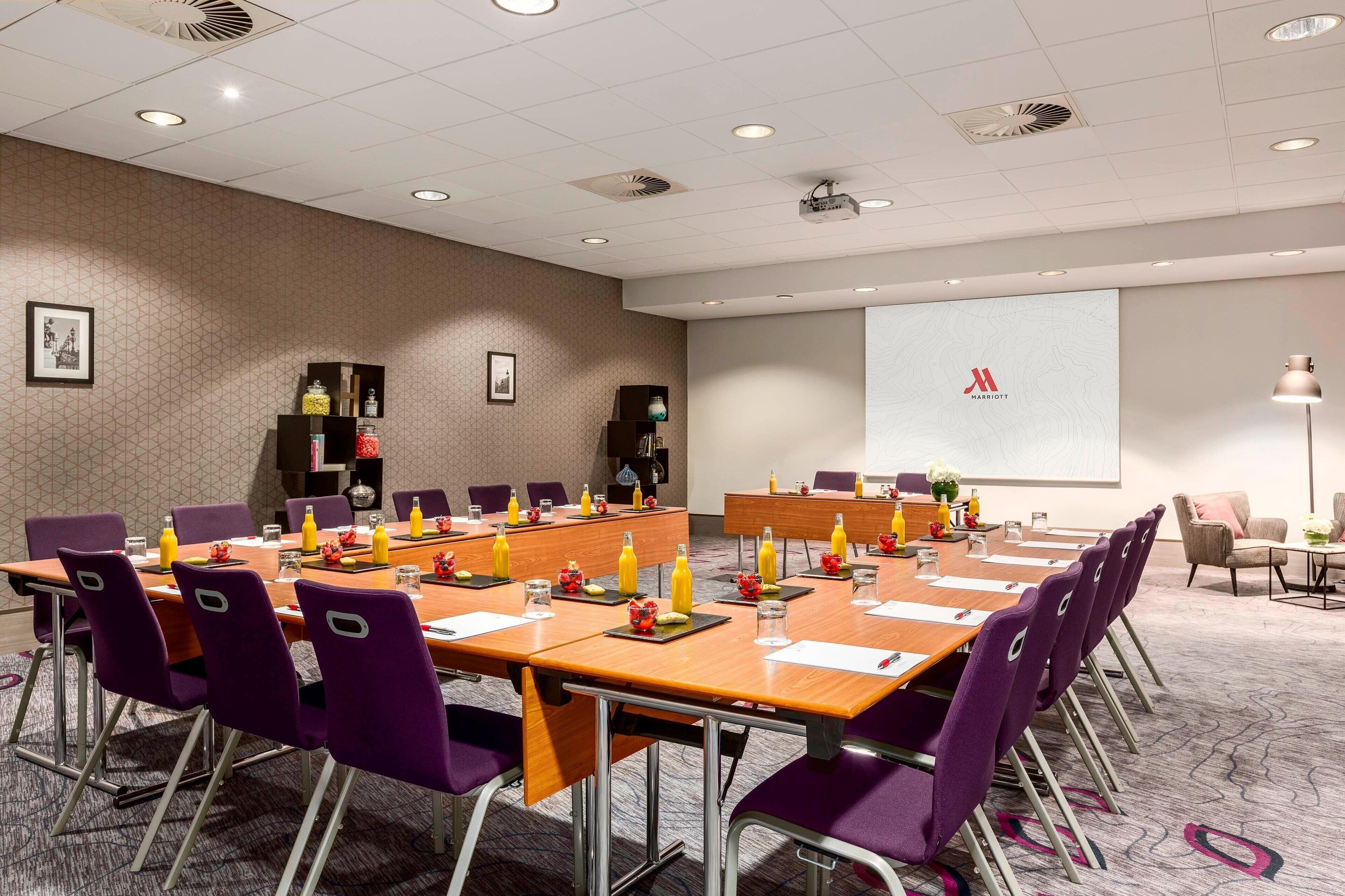 A conference room with a long table, purple chairs, and a projector screen displaying a logo.
