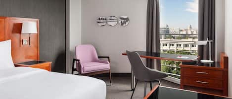Habitación de hotel con cama, escritorio, silla, lámpara, mesita de noche y vista a la ciudad.