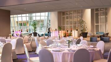 A banquet hall with round tables set for dining, large windows, and indoor plants.