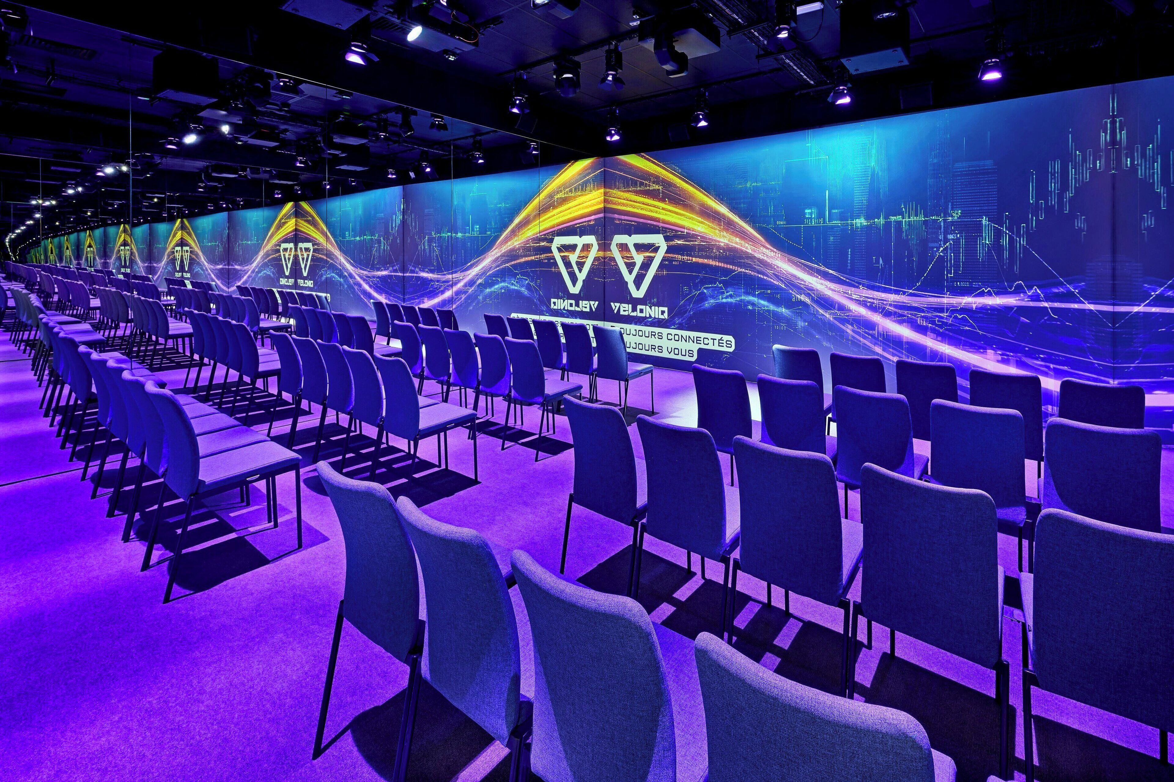 A conference room with rows of chairs and tables, a large digital display with graphs and text, and a ceiling with multiple spotlights.