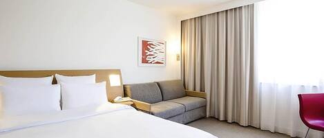 A hotel room with a large bed, a sofa, a red chair, and a window with curtains.