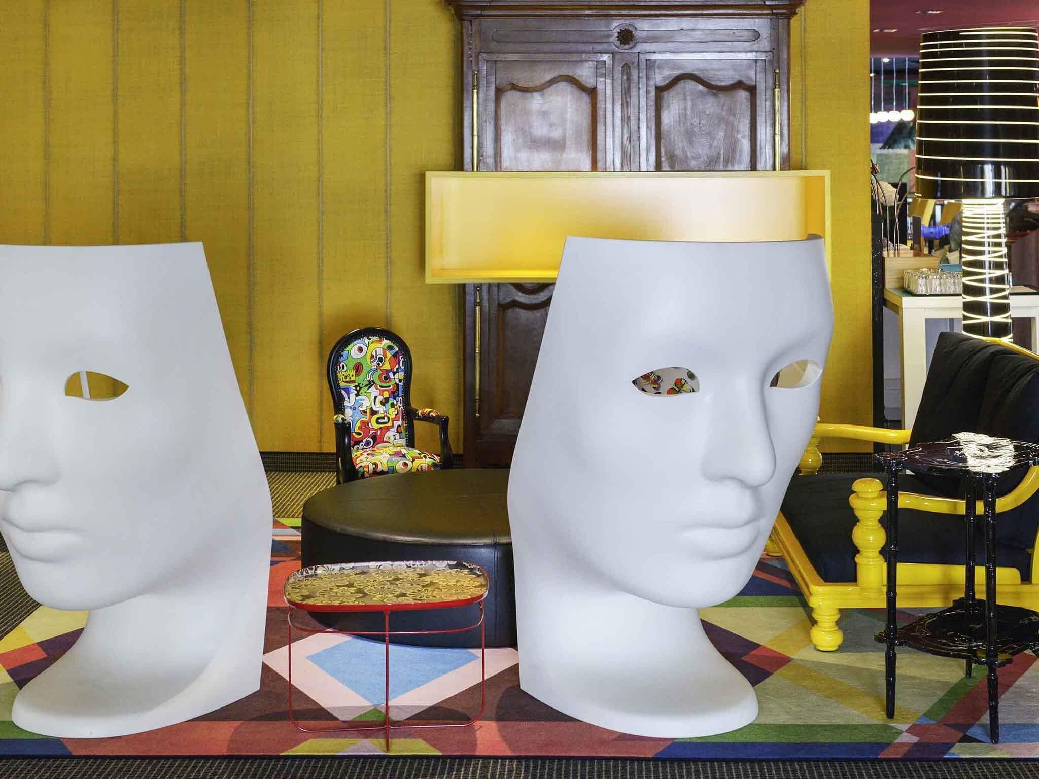 Une pièce comprenant deux grands masques de théâtre blancs, une chaise colorée, une lampe et un fauteuil jaune.