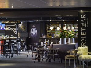 A bar area with a counter, bar stools, hanging lights, and a large screen. - Novotel Metz Centre (Metz)