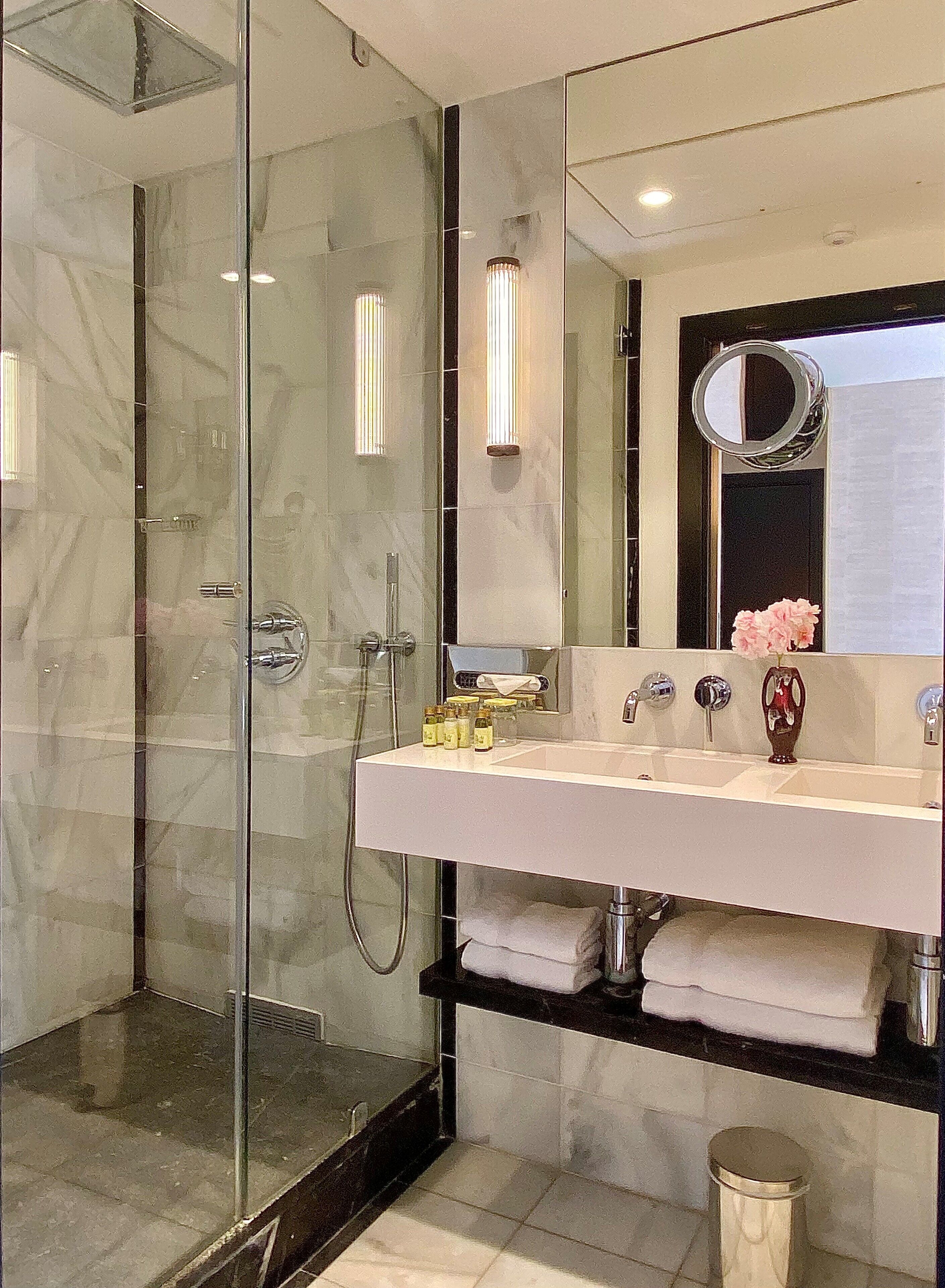 A modern bathroom with a glass-enclosed shower, a large mirror, and a white sink with a vase of flowers.