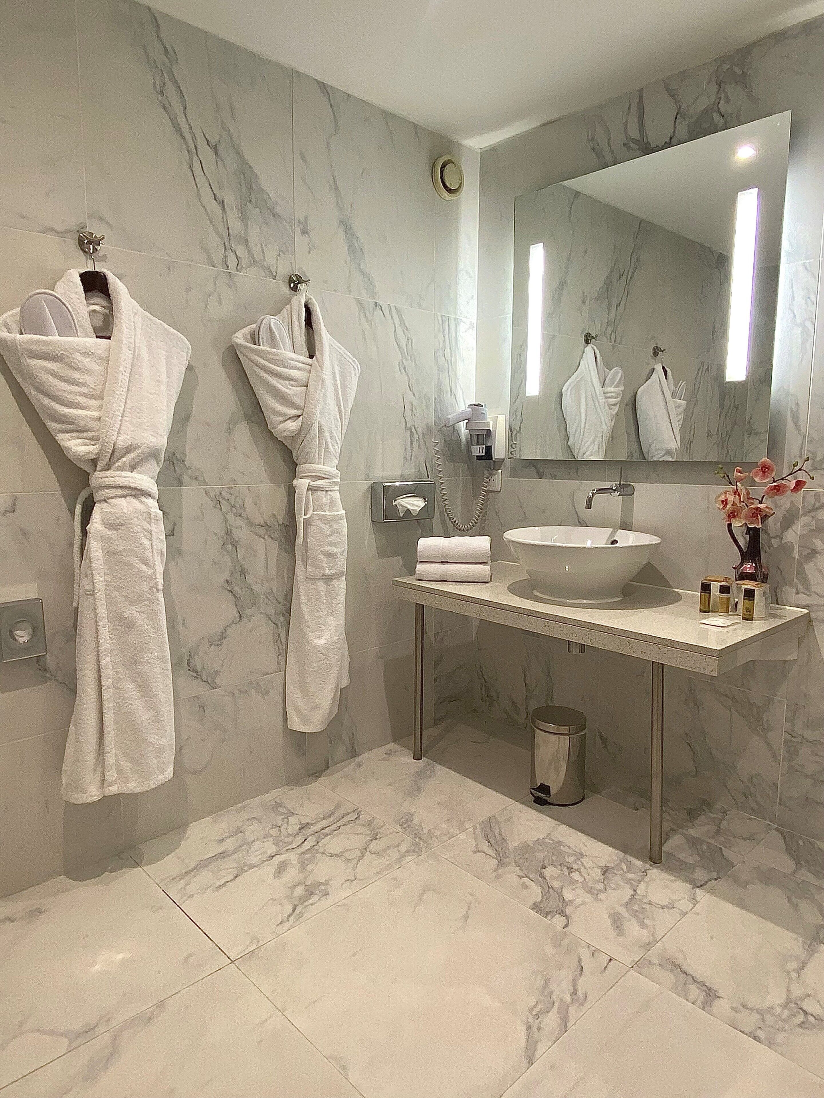 A modern bathroom with marble walls, a large mirror, a sink, and two white bathrobes hanging on hooks.