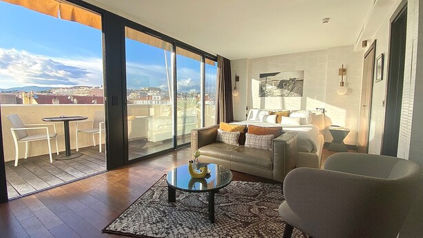 A spacious living room with a large sliding glass door leading to a balcony, a comfortable sofa, a small round table, and a patterned rug. - Hôtel Cannes Centre Univers (Cannes)