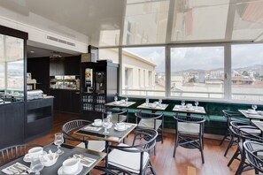 Un restaurante moderno con vistas a la ciudad. Cuenta con una zona de bar, mesas de comedor con sillas y un área de estar con un banco tapizado en verde.