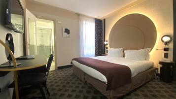 Habitación de hotel con cama, escritorio, silla, espejo, zona de ducha y lámpara fijada a la pared.