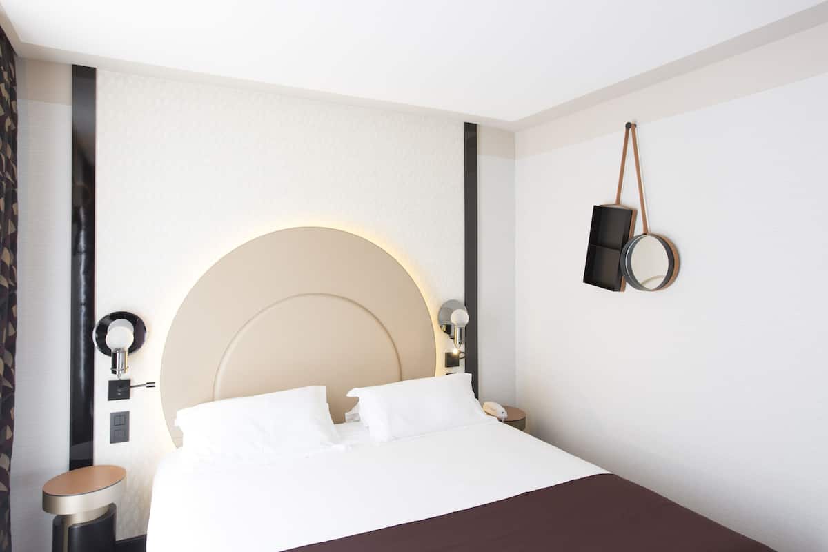A modern hotel room with a large bed, bedside tables, a round mirror, and a small round table.