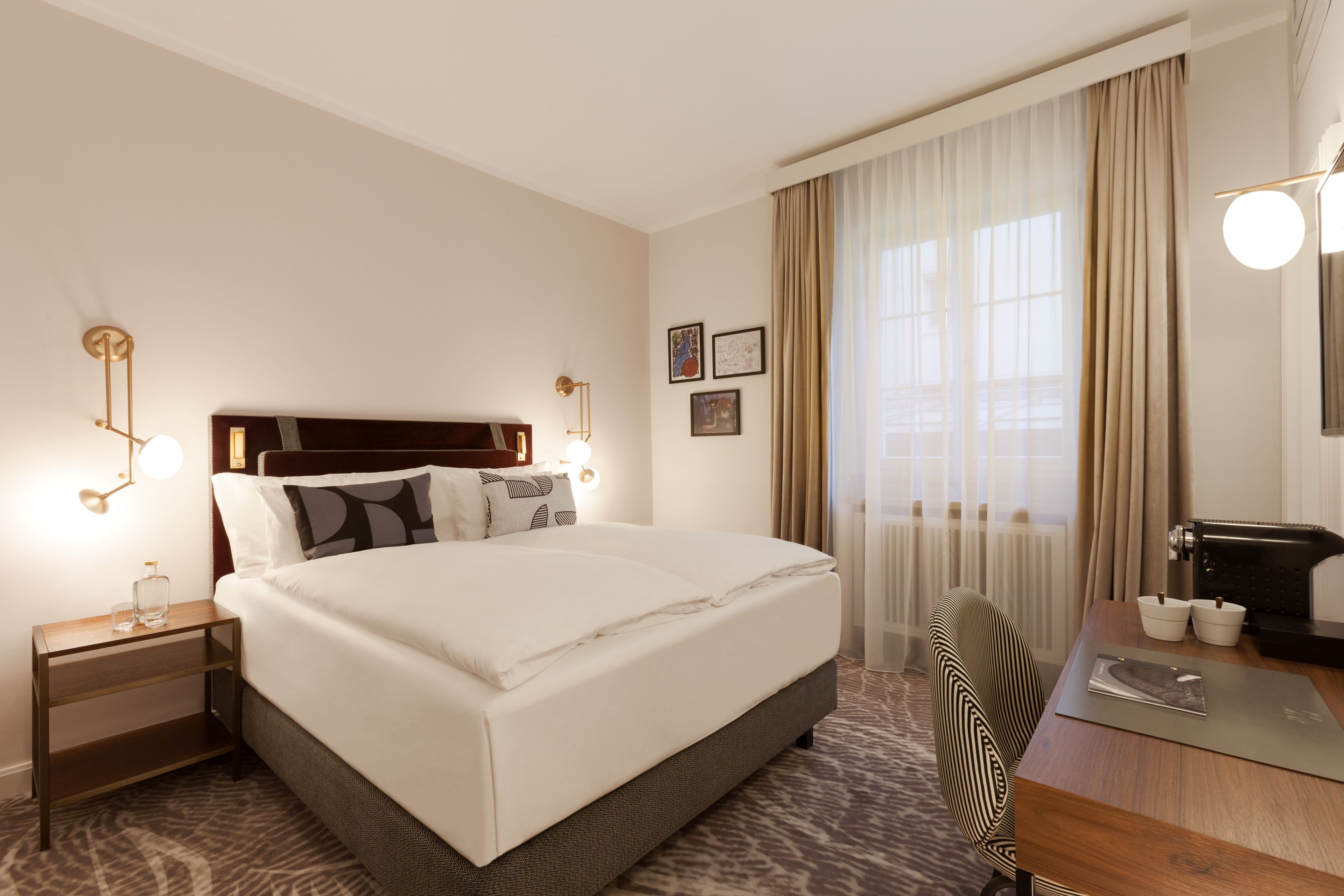 A hotel room with a large bed, bedside tables, a desk with a chair, and a window with curtains.