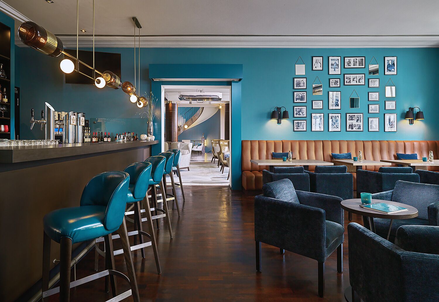 A bar area with a long counter, bar stools, and a seating area with tables and chairs. There are framed pictures on the wall and a staircase leading to another level.