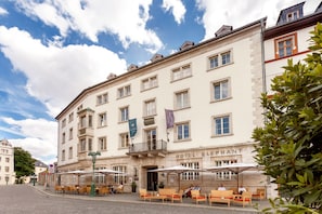 A historic building with a sign reading "HOTEL ELEPHANT." It features a cobblestone street, outdoor seating with umbrellas, and a large tree in the foreground. - Hotel Elephant Weimar, Autograph Collection (Weimar)