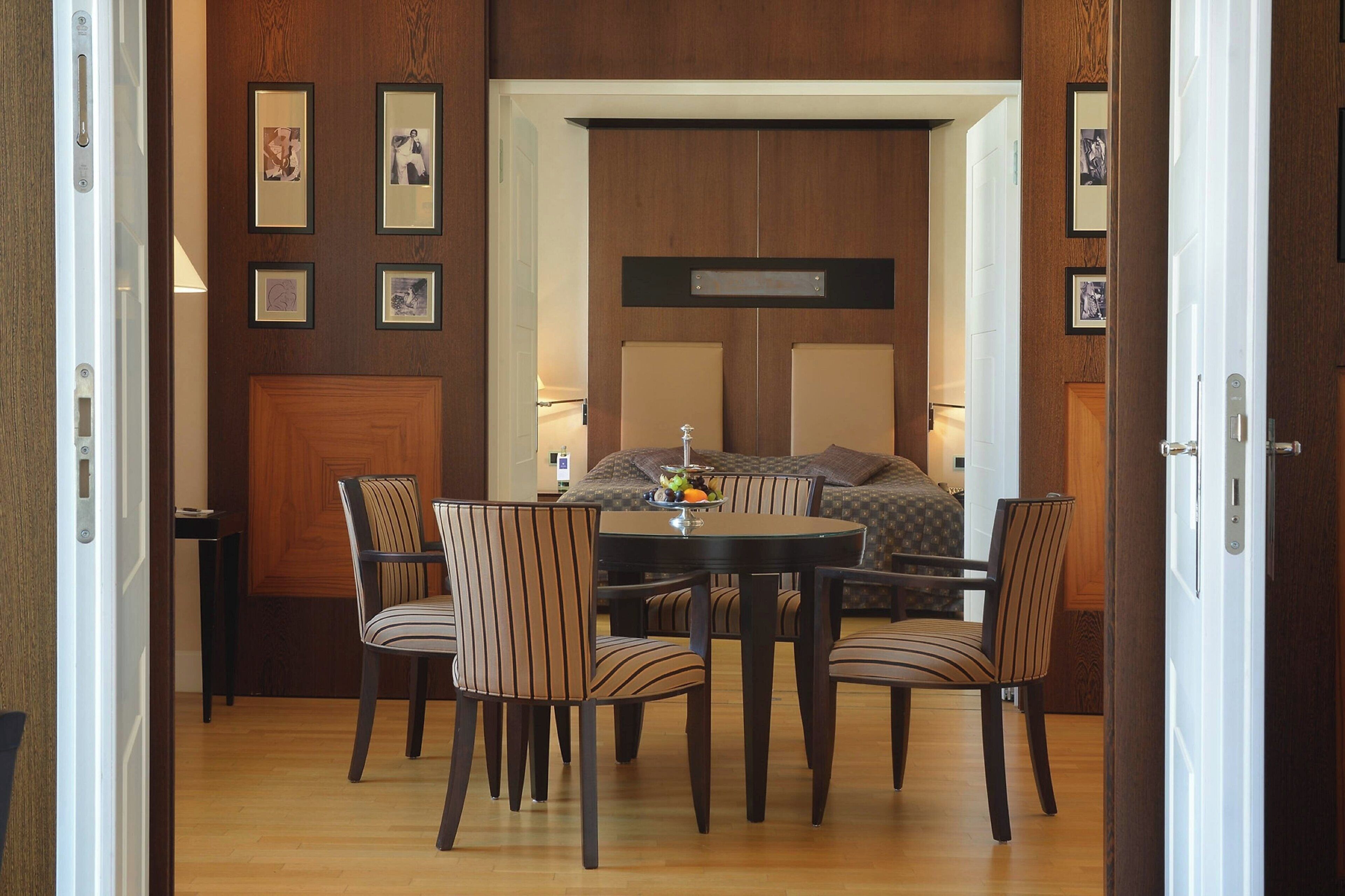 Une chambre d’hôtel avec une porte en bois, un lit doté d’une tête de lit, une table à manger avec des chaises et des tableaux encadrés accrochés au mur.