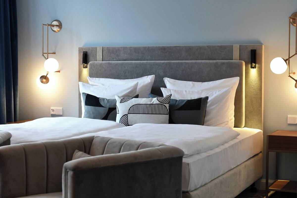 Un lit double avec une tête de lit grise, des oreillers blancs à motifs et des tables de chevet dotées de lampes fixées au mur.
