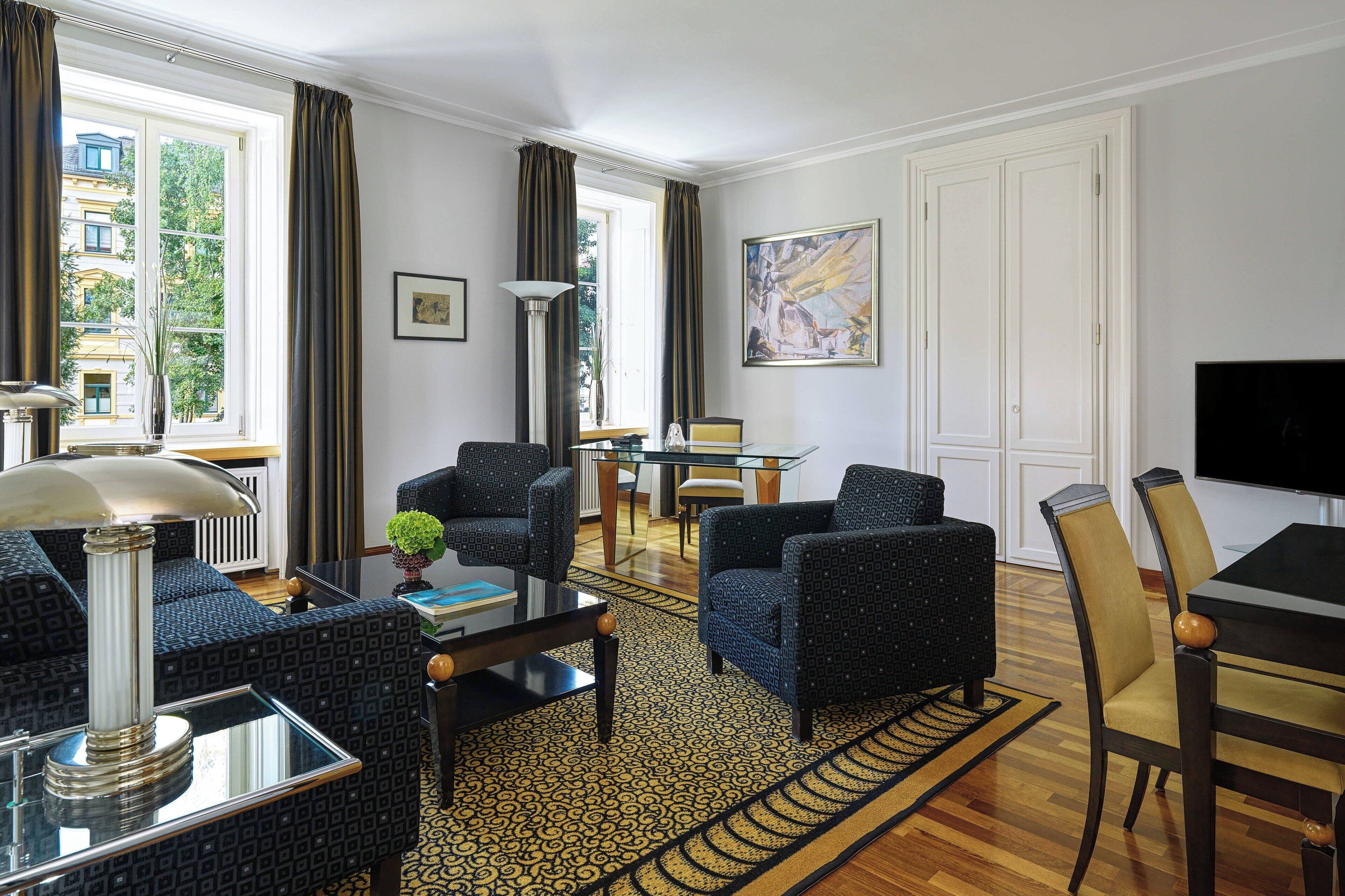 A living room with a sofa, armchairs, a coffee table, and a TV. There is a window with curtains, a painting on the wall, and a lamp.