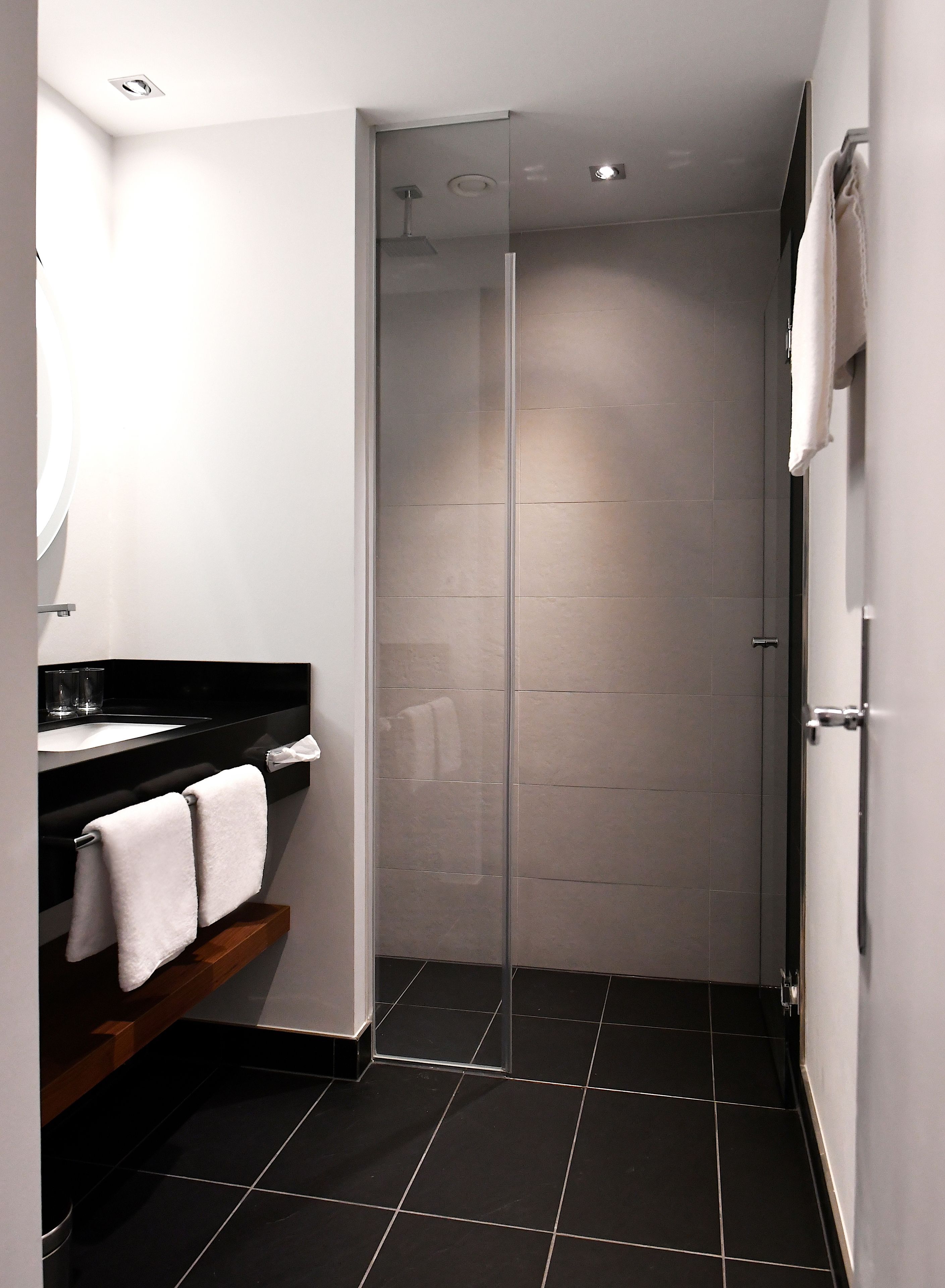 A modern bathroom with a black tiled floor, a glass shower enclosure, a black countertop with a sink, and a wooden shelf with towels.