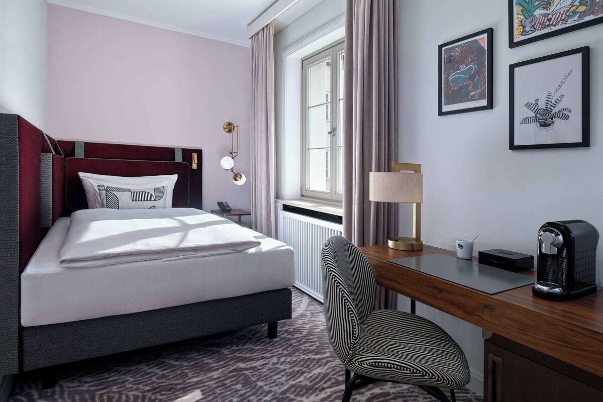 Une chambre d’hôtel équipée d’un lit, d’un bureau, d’une chaise, d’une lampe, d’une machine à café et d’œuvres d’art encadrées accrochées au mur.