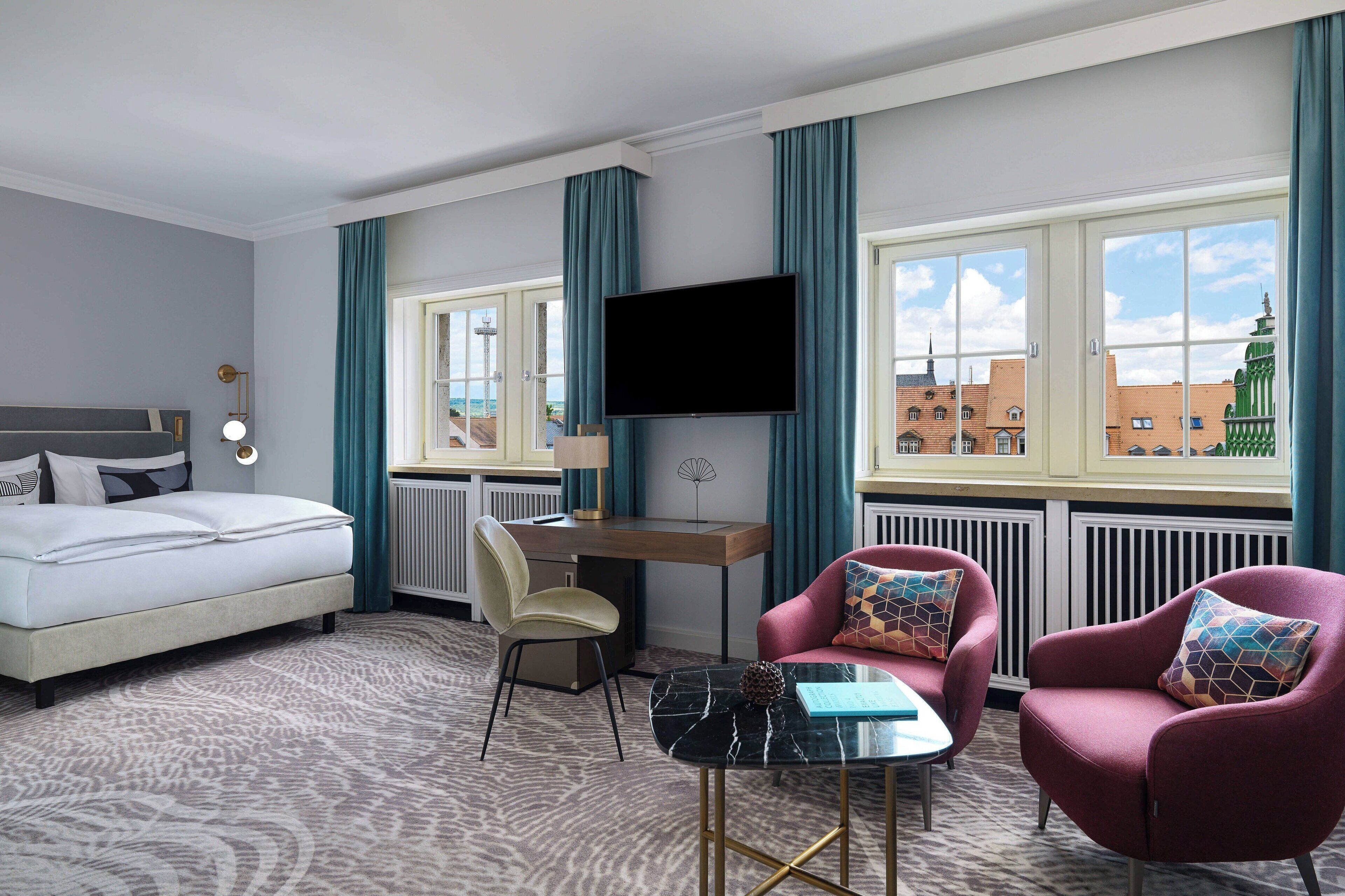 A hotel room with a bed, desk, TV, two armchairs, and a view of buildings through large windows.