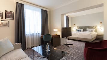 Une chambre d’hôtel avec un canapé, une petite table sur laquelle se trouve une bouteille, une télévision, un lit avec des oreillers et un fauteuil rouge.
