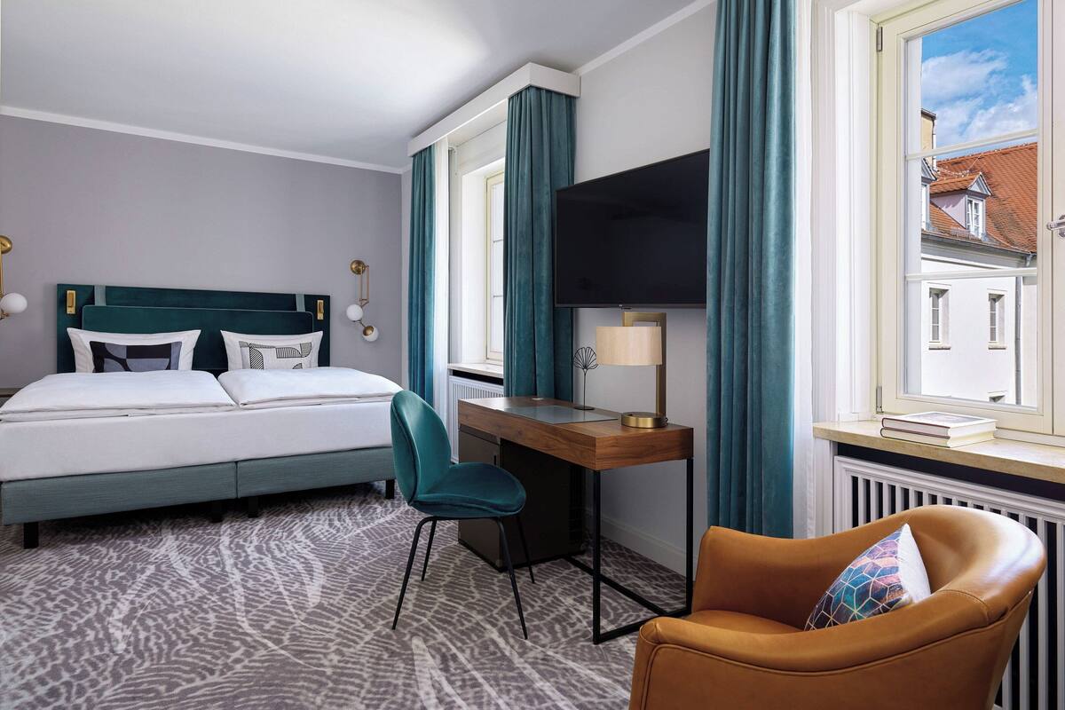 Une chambre d’hôtel avec un lit, un bureau, une chaise et une vue sur les bâtiments, visible par la fenêtre.
